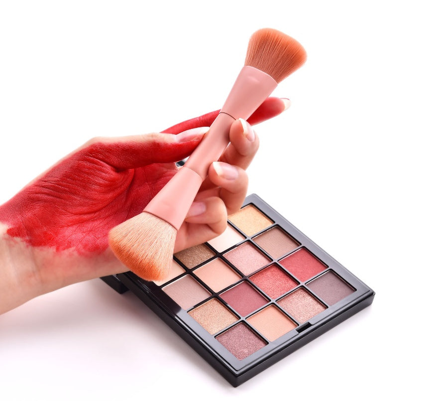 Makeup brush held by a hand with red nail polish next to a makeup palette on a white background