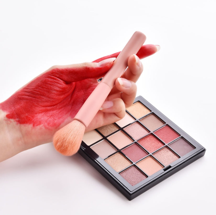 Makeup brush with red makeup stain on hand next to a makeup palette on a white background